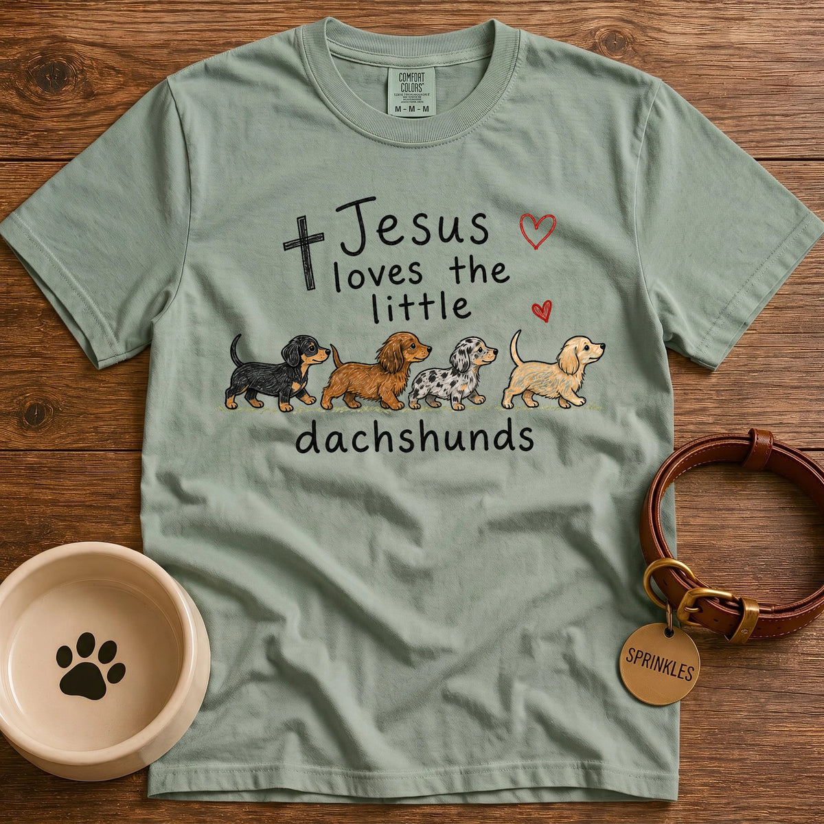 Sage green dachshund T-shirt featuring Jesus Loves the Little Dachshunds lettering, a cross, a heart, and a row of illustrated dachshund dogs.